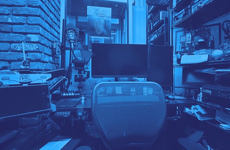View of the office interior: a brick wall, a studio microphone, a teleprompter, two monitors and an empty mesh chair at the desk.